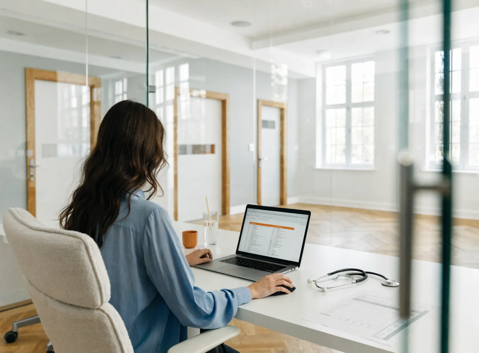 A practice manager at a white desk in a bright clinic office, a laptop showing a search result highlighted at the top